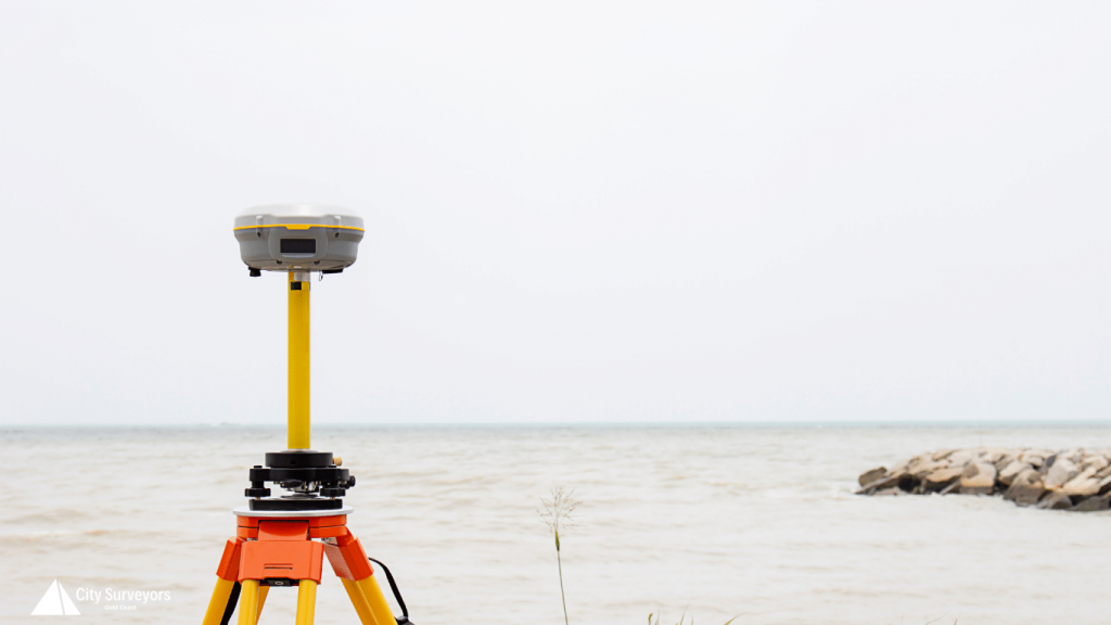 Yellow survey receiver on a tripod beside calm water and rocks, with a small City Surveyors Gold Coast logo in the lower left corner